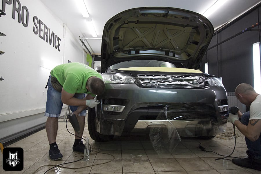 Donde cambiar el aceite del coche uno mismo en Barcelona: Imagen de un taller mecánico con el letrero ‘PRO-SERVICE’ en la pared. Se muestra un vehículo SUV color gris con el capó abierto, situado dentro del taller. Dos personas están trabajando en el coche; una está inclinada hacia la rueda delantera izquierda, posiblemente ajustando o reparando algo, mientras que la otra está agachada al lado derecho del vehículo, realizando tareas de mantenimiento o inspección. El suelo tiene manchas oscuras típicas de un taller mecánico y hay cables dispersos alrededor. La imagen transmite una atmósfera de trabajo y experiencia en servicios automotrices.