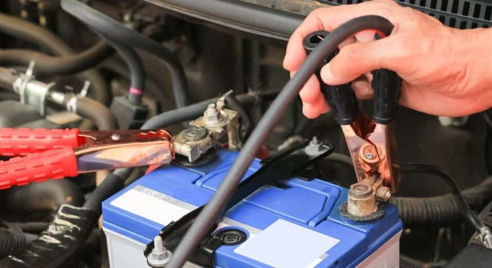 La imagen muestra una batería de coche con cables de arranque conectados. Una mano sostiene la pinza negra, que está conectada al terminal negativo de la batería, mientras que la pinza roja ya está sujeta al terminal positivo. Esta imagen es relevante para demostrar cómo arrancar un coche con pinzas correctamente o para discutir procedimientos de mantenimiento y seguridad del coche.