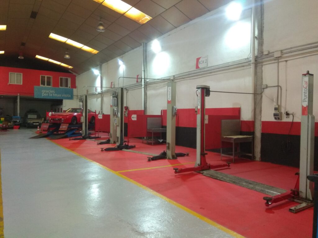 Donde cambiar el aceite del coche uno mismo en Valencia: Imagen de un centro de servicio de vehículos llamado ‘Talleres Puntocar’. Cuenta con varias estaciones de elevación de coches con esquemas de color rojo y blanco, un suelo recubierto de epoxi rojo con líneas de demarcación y una iluminación brillante. La instalación parece limpia, organizada y lista para trabajos de mantenimiento automotriz.