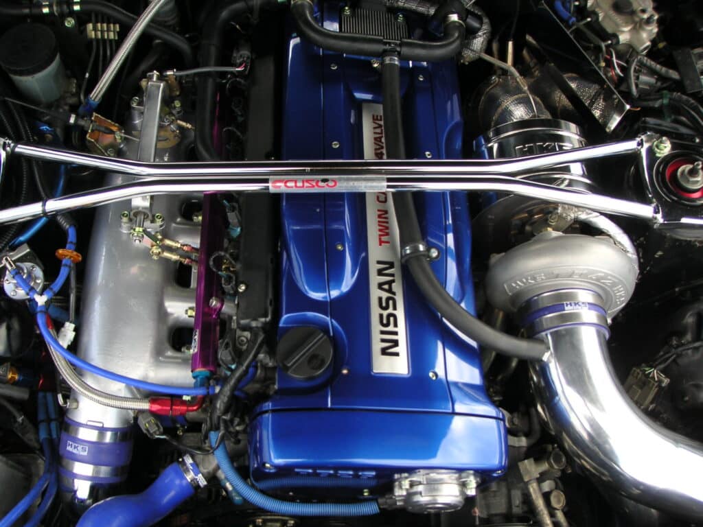 Vista de cerca de un motor RB26DETT del Nissan Skyline R34, pintado de azul con la palabra ‘NISSAN’ destacada. Se observan varios componentes como tuberías, mangueras y partes de la estructura del motor. Una barra plateada con la etiqueta ‘Cusco’ cruza la parte superior del motor. Esta imagen puede ser interesante para aquellos interesados en mecánica automotriz, modificaciones o marcas específicas como Nissan.