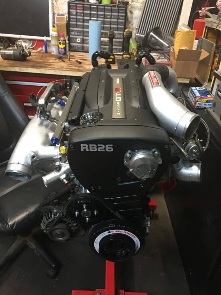 Imagen de un motor RB26DETT de Nissan Skyline. Se observa el motor completo con la tapa de válvulas en color negro y las letras ‘RB26’ en rojo. El motor está montado sobre un soporte rojo, con varias herramientas y cajas visibles al fondo en una estación de trabajo mecánica. Componentes del motor como el turbocompresor, colectores y poleas están expuestos, mostrando su acabado metálico.