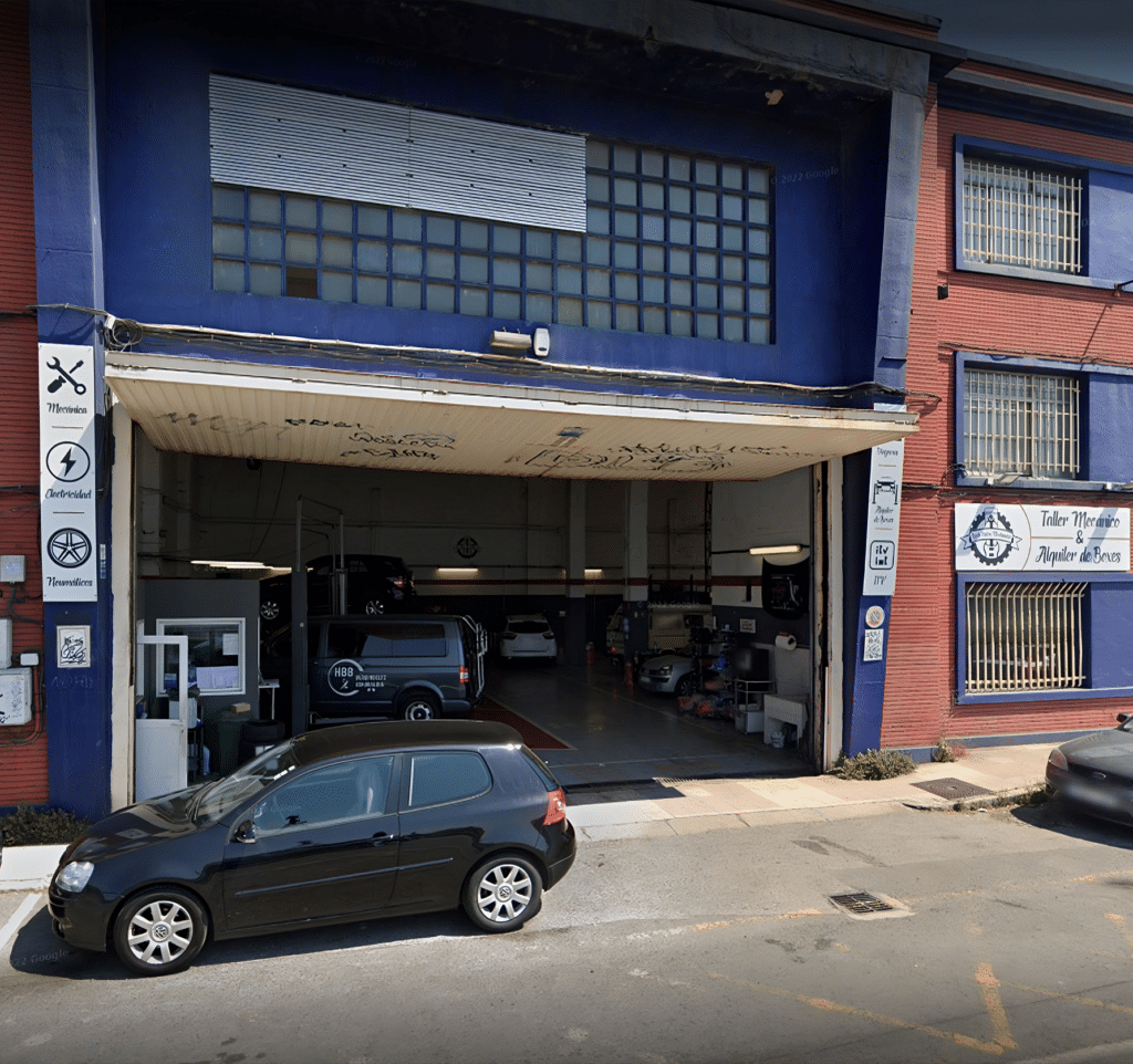 Donde cambiar el aceite del coche uno mismo en Bilbao: Imagen del exterior de un taller de coches con alquiler de boxes llamado ‘Rock Piston Mechanics’. El edificio de dos pisos tiene una fachada azul y roja, con un garaje en la planta baja. Hay vehículos estacionados dentro y fuera del garaje, incluyendo un coche negro frente a la entrada. El edificio tiene letreros que indican que es un lugar de servicio automotriz, con referencias específicas a ‘Rock Piston Mechanics’ y ‘Tire World’.