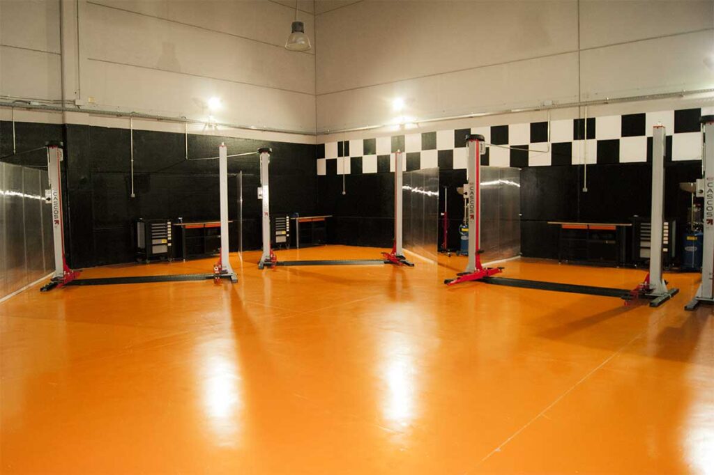 Dónde cambiar el aceite del coche uno mismo en Madrid: Vista interior de las instalaciones de RTMBOX, un taller mecánico vacío con suelo naranja brillante. Cuatro elevadores de vehículos blancos y rojos están uniformemente distribuidos en el espacio. Las paredes son negras con detalles decorativos en blanco y negro que recuerdan a la bandera a cuadros de una carrera, lo que sugiere un enfoque en la velocidad y el rendimiento automotriz. No hay personas ni vehículos presentes, lo que indica una imagen tomada posiblemente fuera del horario laboral o para fines promocionales.