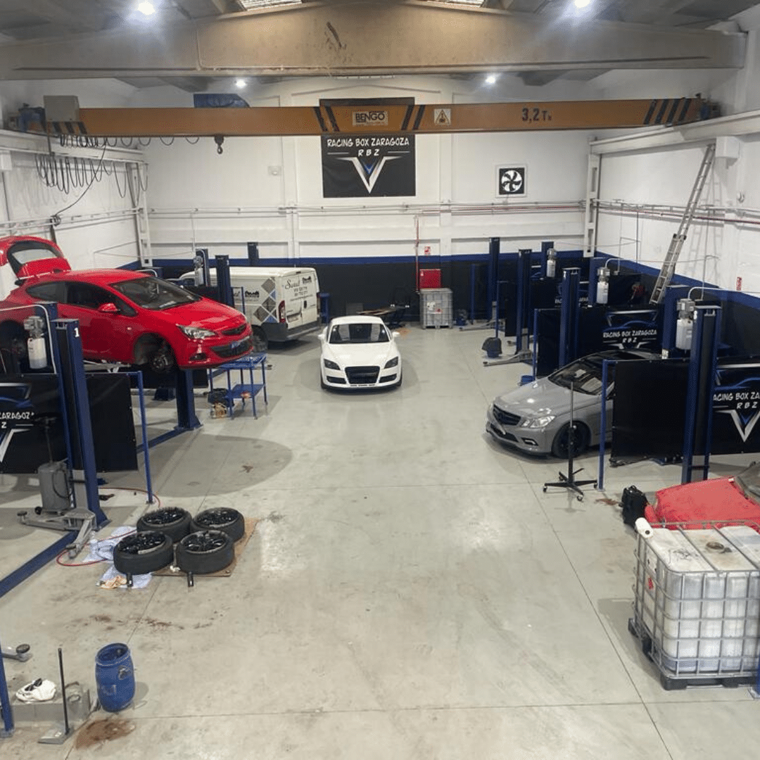 Interior de Racing Box Zaragoza, un taller de alquiler de boxes donde cambiar el aceite del coche uno mismo en Zaragoza. Se observan varios coches siendo atendidos, uno rojo en un elevador y otro blanco en el suelo. El espacio es amplio y cuenta con diversas herramientas y maquinaria. Un cartel con el nombre ‘RACING BOX ZARAGOZA’ es visible al fondo.