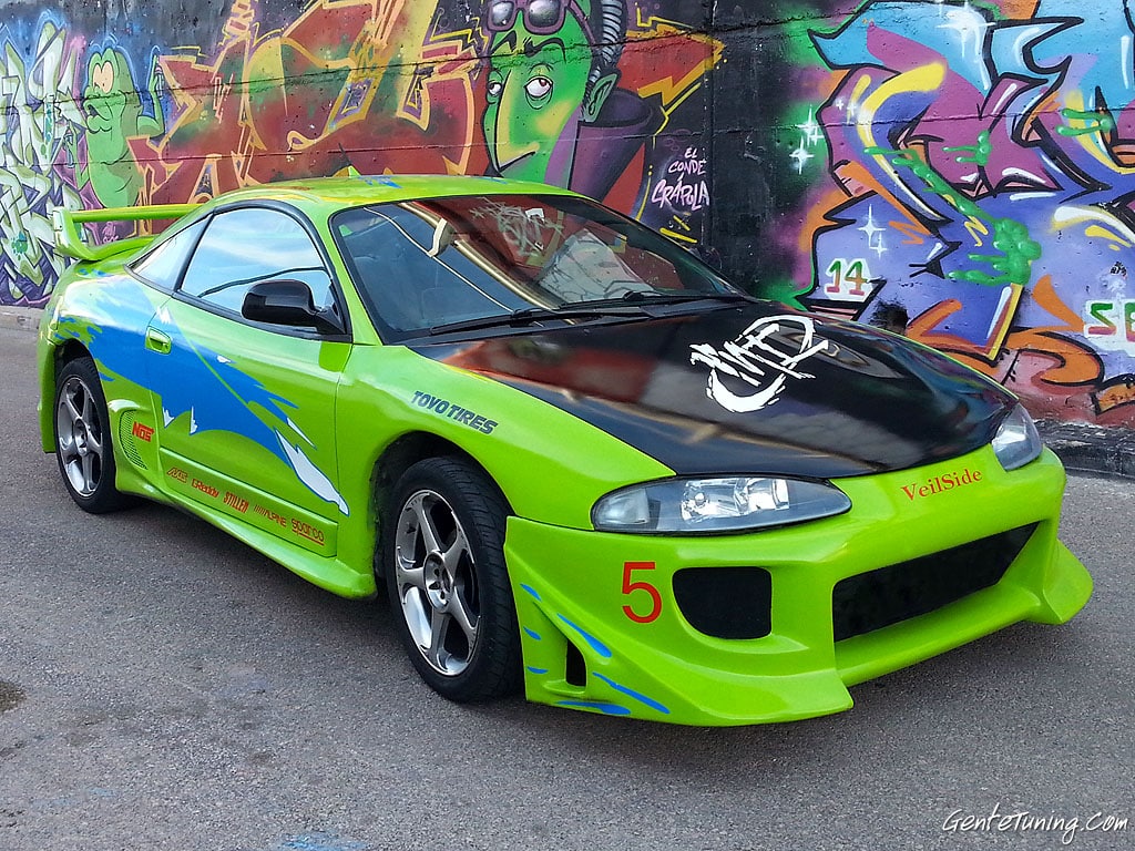 Coche deportivo verde brillante con gráficos azules y plateados en el lateral, similar al Mitsubishi Eclipse de la película ‘Fast and Furious’. El coche está estacionado frente a una pared con arte de graffiti colorido. Tiene el número ‘5’ en la puerta y modificaciones como un kit de carrocería, alerón trasero y ruedas personalizadas.