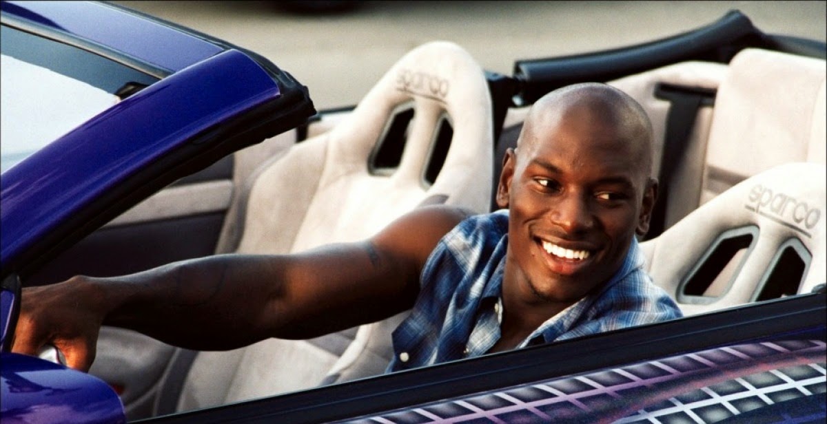 Roman Pearce (Tyrese Gibson) sentado en el asiento del conductor de un coche descapotable con exterior púrpura e interior blanco. Los reposacabezas tienen la marca ‘SPARCO’. Roman lleva una camisa de cuadros y está extendiendo un brazo, posiblemente para abrir o cerrar la puerta. La imagen está asociada con la franquicia ‘The Fast and the Furious’.