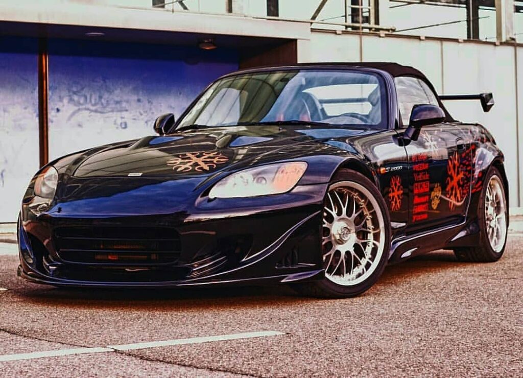 Honda S2000 de Fast and Furious 1, negro con gráficos personalizados, estacionado. El vehículo presenta una pintura negra brillante con distintivas calcomanías de llamas naranjas y rojas en los laterales y el capó. Tiene un kit de carrocería deportivo, incluyendo un parachoques delantero con grandes entradas de aire y faldones laterales. El coche está equipado con llantas blancas de múltiples radios y parece estar en perfectas condiciones, reflejando su papel como el coche de Johnny Tran en la película.