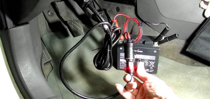 Un salvamemorias de coche conectado al puerto OBD de un vehículo, con cables conectados a una batería portátil. Una mano sostiene la pinza roja, lista para conectarla a la batería del coche.