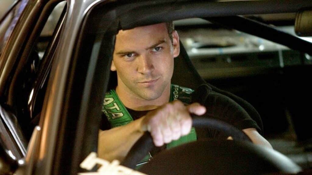 Sean Boswell (Lucas Black) sentado en el asiento del conductor de un coche, sosteniendo el volante con ambas manos. Lleva una camiseta verde y parece estar concentrado en algo fuera del vehículo. La imagen está asociada con la franquicia ‘The Fast and the Furious’.
