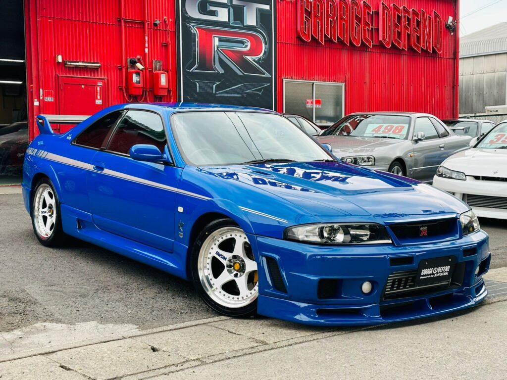 Un Nissan Skyline GT-R R33 azul estacionado frente a un edificio con el letrero ‘GT-R GARAGE DEFEND’. El vehículo cuenta con llantas blancas de carreras, un kit de carrocería de posventa con parachoques delantero y faldones laterales, y una ventilación prominente en el capó. El coche está posicionado para mostrar su perfil, destacando el diseño aerodinámico y la postura deportiva que lo hacen interesante para los entusiastas de los coches y relevante en discusiones sobre vehículos de alto rendimiento.