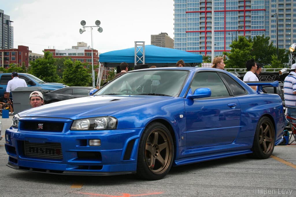 Un Nissan Skyline GT-R R34 azul estacionado en un evento al aire libre con varias personas y una carpa en el fondo. El coche destaca por su diseño deportivo, con una prominente toma de aire en el capó, líneas aerodinámicas y llantas de bronce de posventa que sugieren mejoras de rendimiento. Este modelo es popular entre los entusiastas del automovilismo debido a su historia en las carreras y su estatus como un icónico coche deportivo japonés.