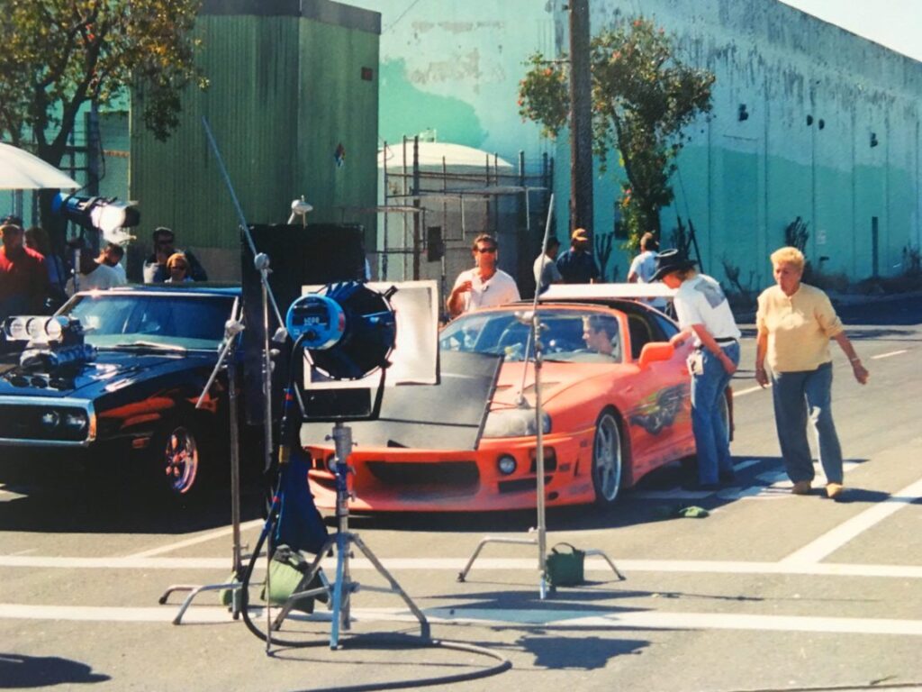 Imagen del rodaje de ‘Fast and Furious 1’ (2001) mostrando dos coches icónicos: un Dodge Charger negro y un Toyota Supra naranja. La escena se desarrolla en un entorno urbano al aire libre, con equipo de producción como cámaras y luces, y miembros del equipo de filmación alrededor. Es coche de Brian en Fast & Furious.