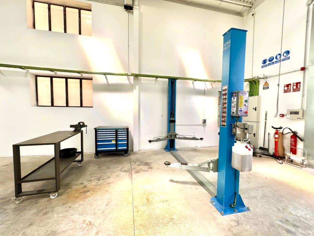 Donde cambiar el aceite del coche uno mismo en Barcelona: Imagen de un espacio de trabajo compartido identificado como ‘The CoMaking Space’. Se observan diversas herramientas y equipos típicos para la construcción o reparación de objetos. En el centro hay un elevador de coches azul, una mesa de trabajo metálica a la izquierda y unidades de almacenamiento con cajones probablemente para herramientas. A la derecha, hay equipo de seguridad, incluyendo un extintor. El espacio tiene ventanas de estilo industrial en la parte alta de la pared y una iluminación brillante. La imagen es relevante para aquellos interesados en proyectos de bricolaje, mecánica o espacios de trabajo compartidos.