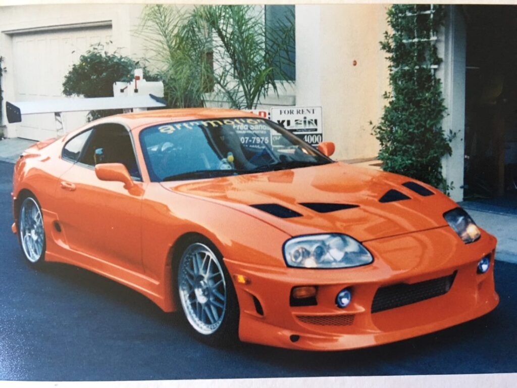 Toyota Supra naranja de ‘Fast and Furious 1’ (2001) estacionado en una calle residencial antes de ser personalizado con vinilos. El coche presenta un diseño deportivo con una prominente toma de aire en el capó y un kit aerodinámico.