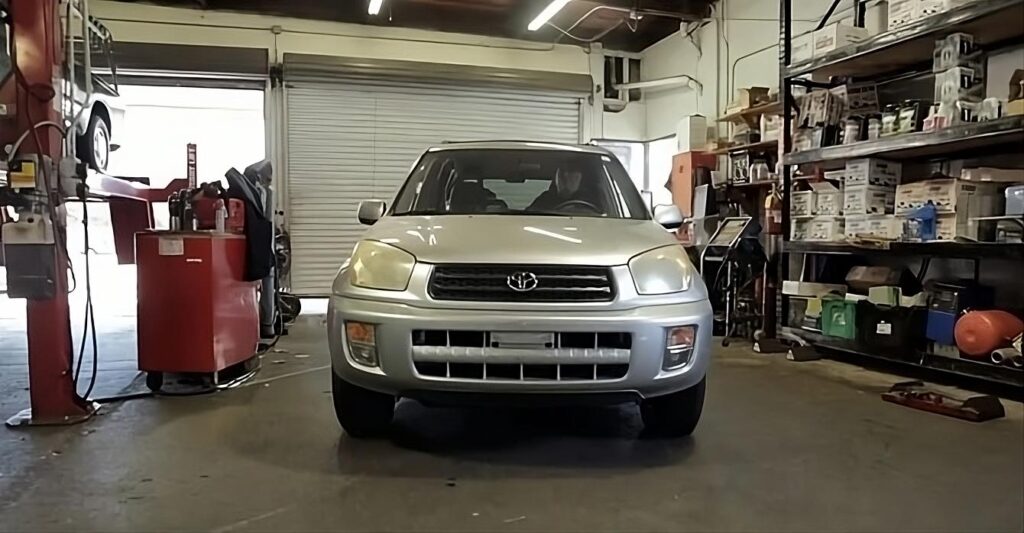 Imagen de un Toyota RAV4 plateado estacionado en un garaje espacioso con varias herramientas y equipos visibles en el fondo. El vehículo está posicionado sobre una superficie plana de hormigón que se extiende por toda el área del garaje. El capó del coche está cerrado y el garaje tiene una atmósfera abierta con luz natural entrando desde el exterior.