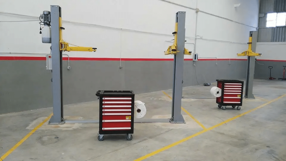 Donde cambiar el aceite del coche uno mismo en Valencia: Imagen de un espacio de mantenimiento de coches de autoservicio llamado ‘Tu Box Mecánica’. Hay dos elevadores de vehículos amarillos instalados en el suelo, utilizados para levantar coches y trabajar debajo de ellos. A la derecha, hay un cofre de herramientas rojo y blanco con múltiples cajones, y junto a él, un carrete blanco posiblemente para fluidos o aire. El suelo es gris con una línea roja marcando la parte inferior de las paredes. Este entorno es relevante para alguien que busca cambiar el aceite de su coche por sí mismo en Valencia, ya que proporciona el equipo y el espacio necesarios para hacerlo.