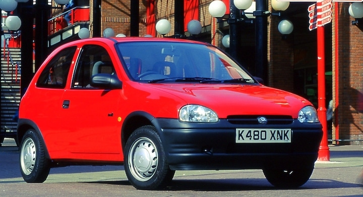 Un Opel Corsa rojo está estacionado en una calle durante el Festival of the Unexceptional. El coche está orientado ligeramente hacia la izquierda del encuadre, mostrando su perfil frontal y lateral. En el fondo, se ve un cielo despejado con algunos edificios y lo que parece ser una farola. La matrícula del coche es visible.