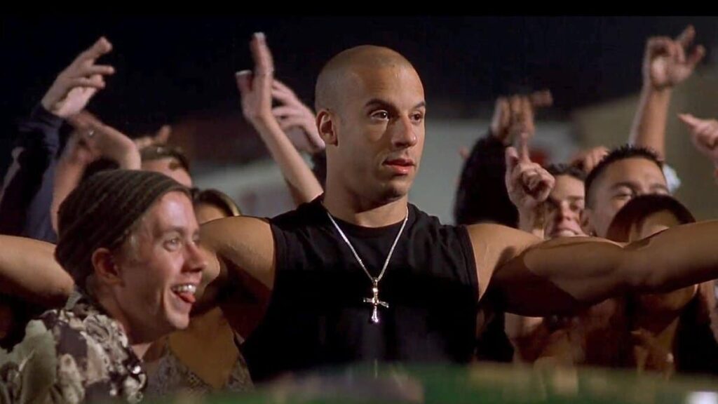 Vin Diesel de frente, con una camiseta sin mangas negra y un collar plateado, en una escena nocturna al aire libre. Varias personas con los brazos levantados lo rodean. La imagen está asociada con la franquicia ‘The Fast and the Furious’.