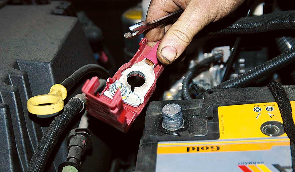 Primer plano de una mano usando una llave para aflojar o apretar el perno en el terminal de una batería de coche. La batería tiene una etiqueta amarilla con texto parcialmente visible y varios cables y componentes del vehículo alrededor.