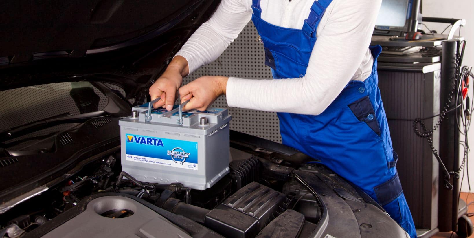 La imagen muestra a una persona con un uniforme azul y blanco, posiblemente un mecánico, instalando una batería de coche de la marca VARTA en el compartimento del motor de un vehículo. El enfoque está en el proceso de instalación de la batería, lo cual es relevante para aquellos interesados en el mantenimiento automotriz o en aprender sobre los componentes del vehículo.
