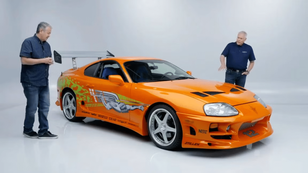 Craig Lieberman junto a un Toyota Supra naranja con calcomanías y un gran alerón trasero, conocido por su aparición en la película ‘The Fast and the Furious’. La imagen está tomada en un estudio con fondo blanco.