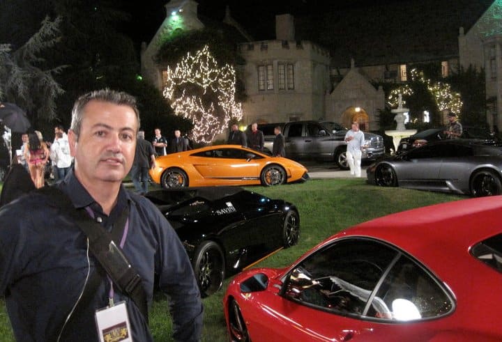 Reunión de personas en un evento al aire libre durante la noche, con árboles iluminados y un gran edificio al fondo. En primer plano, tres coches deportivos: uno naranja, uno negro y uno rojo. La imagen muestra un evento de exhibición de coches de lujo, con Craig Lieberman presente.