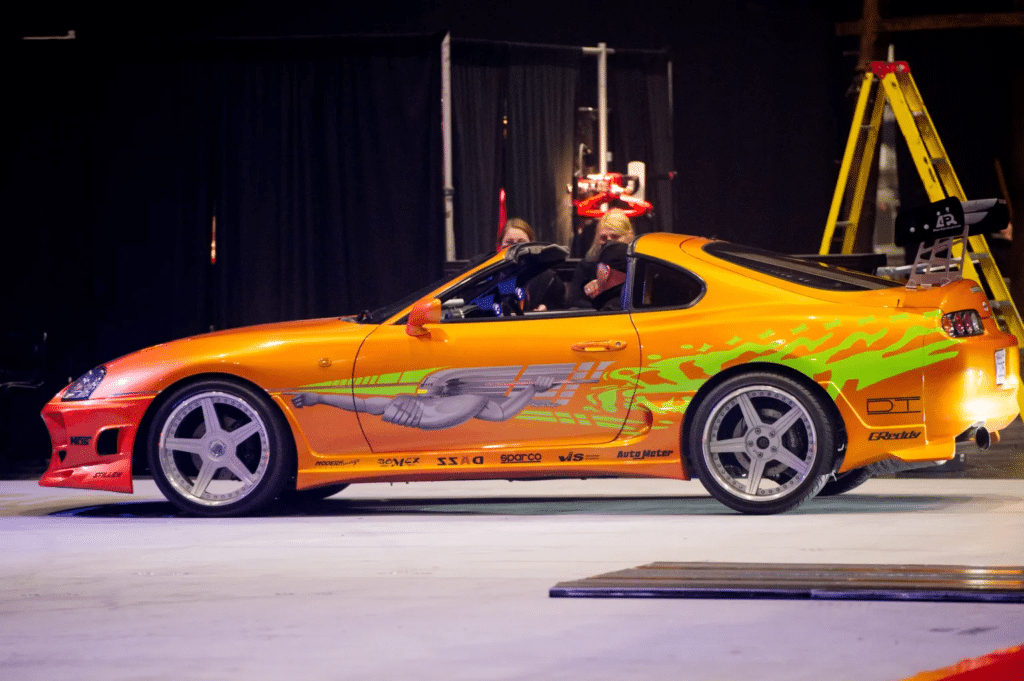 Vista lateral del Toyota Supra naranja de ‘Fast and Furious 1’ (2001) con calcomanías gráficas, incluyendo llamas y un diseño de gladiador, en un entorno interior con iluminación artificial. Hay una persona visible dentro del coche y otra de pie junto al vehículo. En el fondo, hay un equipo que parece ser una grúa o dispositivo de elevación.