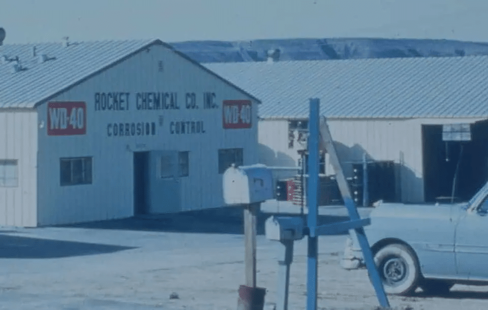 Imagen histórica de la fábrica de WD-40 Multiusos en sus inicios. El edificio industrial tiene un cartel que dice “ROCKET CHEMICAL CO. WD-40 CORROSION CONTROL”. En primer plano, hay un buzón y un coche estacionado a la derecha. Esta imagen representa los comienzos de la marca WD-40 Multiusos.