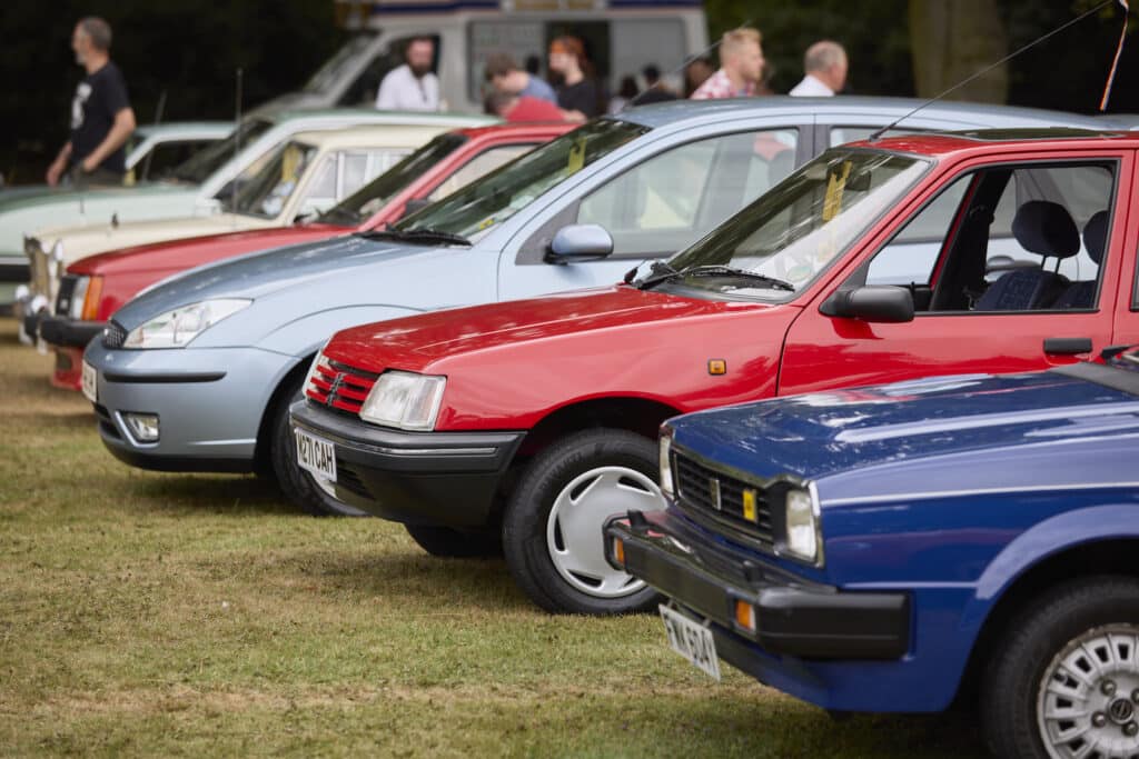 Una fila de cuatro coches está estacionada sobre el césped en lo que parece ser un festival de coches. Los vehículos están en primer plano, con personas de pie y caminando en el fondo. Los coches están orientados de manera que sus frentes miran ligeramente hacia la cámara, mostrando sus diseños y colores. De más cerca a más lejos, hay un Honda Civic de los 70, color azul, un Peugeot 205 rojo, un Ford Focus mk1 plateado y un Opel Corsa A de color rojo. Esta imagen captura un momento del Festival of the Unexceptional, celebrando el diseño y la historia de estos vehículos.