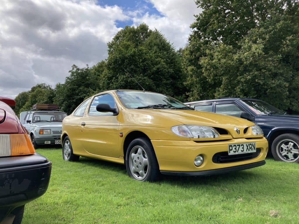 Un Renault Megane amarillo mostaza con matrícula P373 XRM está estacionado sobre el césped en un evento al aire libre, presumiblemente el Festival of the Unexceptional. El coche, un modelo de dos puertas con los faros encendidos, está en primer plano. En el fondo, se ven otros coches y árboles bajo un cielo nublado, sugiriendo un día gris.