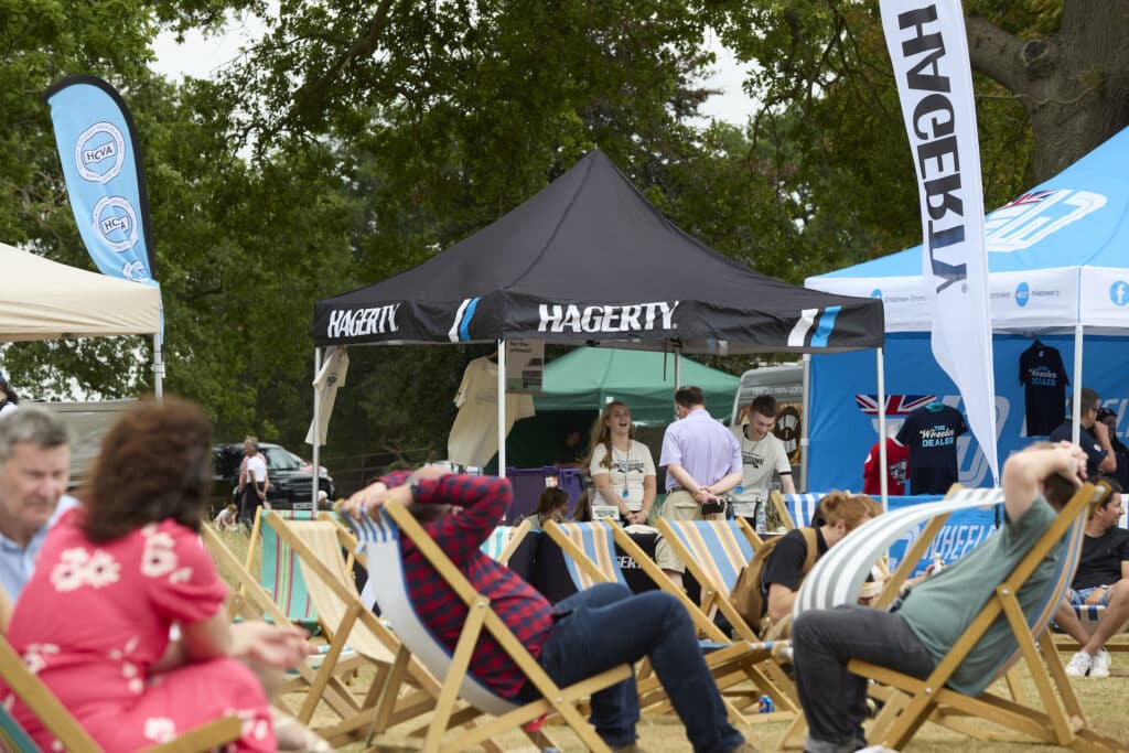 Varias personas están sentadas en sillas de madera al aire libre, mirando hacia una carpa negra con la palabra ‘HAGERTY’ impresa. También hay banderas azules con logotipos, uno de los cuales parece ser el de Ford. El entorno sugiere una reunión social casual, posiblemente relacionada con intereses automovilísticos, durante el Festival of the Unexceptional.