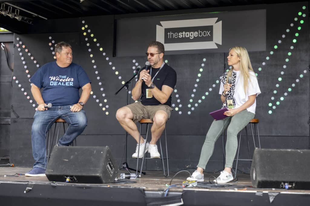 Tres personas están en un escenario con micrófonos, presumiblemente participando en una charla o presentación durante el Festival of the Unexceptional. El fondo presenta un cartel que dice ‘thestagebox’ y otro con el texto ‘The Wheeler Dealer’, relacionado con el programa de televisión ‘Wheeler Dealers’. Entre los participantes está Mike Brewer, conocido presentador de ‘Joyas sobre ruedas’.