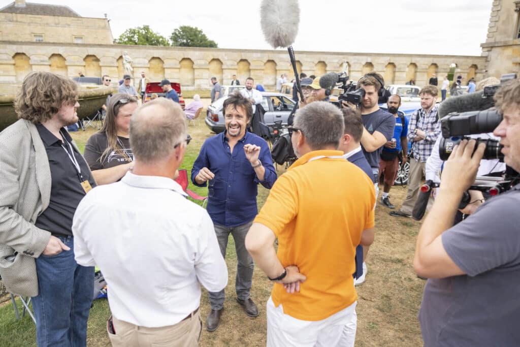 Un grupo de personas está reunido alrededor de Richard Hammond, presentador de ‘Top Gear’, quien está hablando con los asistentes durante el Festival of the Unexceptional. El evento se lleva a cabo al aire libre en Grimsthorpe Castle, y hay varias cámaras y micrófonos apuntando hacia él.
