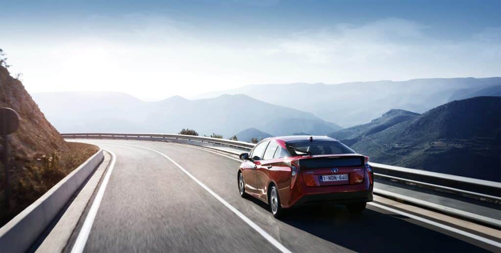 Imagen de un Toyota Prius de última generación, de color rojo, conduciendo por una autopista con un paisaje escénico de montañas y cielo despejado de fondo. El vehículo está en el carril derecho y la carretera se curva suavemente hacia la izquierda. La iluminación sugiere que podría ser temprano en la mañana o al final de la tarde.