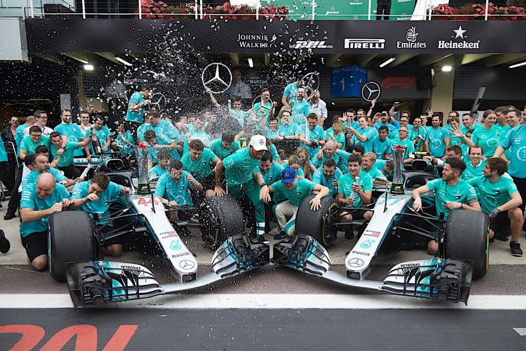 Escena de celebración con dos coches de Fórmula 1 de Mercedes-Benz y un gran grupo de miembros del equipo con camisetas turquesas. El equipo está reunido en una pista de carreras, y varias personas están rociando lo que parece ser champán, creando un ambiente festivo. Esta imagen captura el momento de victoria y celebración del equipo Mercedes-Benz en un evento de Fórmula 1, probablemente significando una victoria importante o un título de campeonato en 2018.