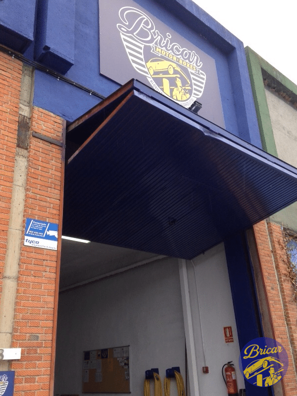 Donde cambiar el aceite del coche uno mismo en Madrid: Imagen de la fachada de un establecimiento llamado ‘Bricar’, con un logotipo que incluye un escudo y dos llaves cruzadas. La entrada tiene una marquesina azul y hay un letrero azul con el nombre ‘Bricar’ en letras blancas. A la izquierda, se observa una señalización vertical que dice ‘Taller’ con una flecha apuntando hacia la derecha, indicando la dirección del taller.