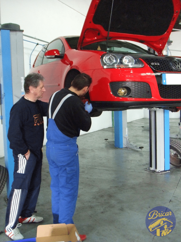 Donde cambiar el aceite del coche uno mismo en Madrid: Imagen de dos personas en un taller de mantenimiento de vehículos. Una persona, vestida con un mono azul, está trabajando en un coche rojo levantado del suelo, posiblemente inspeccionándolo o reparándolo. La otra persona está de pie cerca, observando el trabajo. Las caras de ambas personas están ocultas por privacidad.
