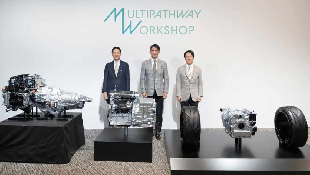 Tres motores en exhibición con tres figuras detrás de ellos, con el texto ‘Multipathway Workshop’ arriba. Representa la colaboración entre Toyota, Subaru y Mazda para el desarrollo de motores de combustión eficientes y compatibles con combustibles neutros en carbono.