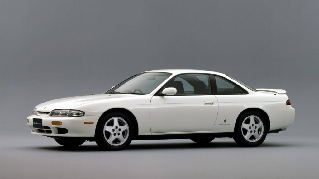 Imagen de un Nissan Silvia S14 de color blanco, un coupé deportivo de dos puertas, mostrado contra un fondo gris liso. El vehículo se ve desde un ángulo lateral-frontal, destacando su diseño elegante y líneas suaves. El coche cuenta con faros escamoteables, una parrilla frontal distintiva y llantas de aleación de cinco radios. No es un Nissan Silvia 2028