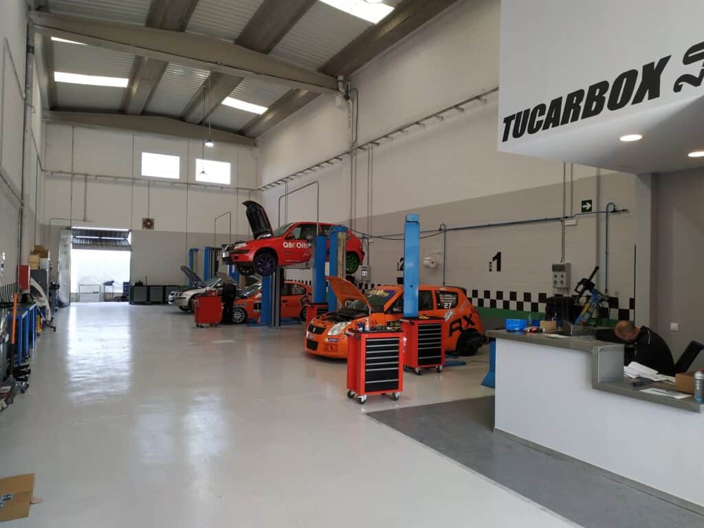 Donde cambiar el aceite del coche uno mismo en Madrid: Imagen del interior de un taller de servicio de vehículos. Hay dos elevadores hidráulicos con vehículos levantados, varios cofres de herramientas y equipos para el mantenimiento de coches. En primer plano, se ve un mostrador de recepción con una persona sentada detrás. El taller es espacioso y bien iluminado, con grandes ventanas o tragaluces que permiten la entrada de luz natural. El nombre ‘TUCARBOX’ se muestra de manera prominente en la pared.