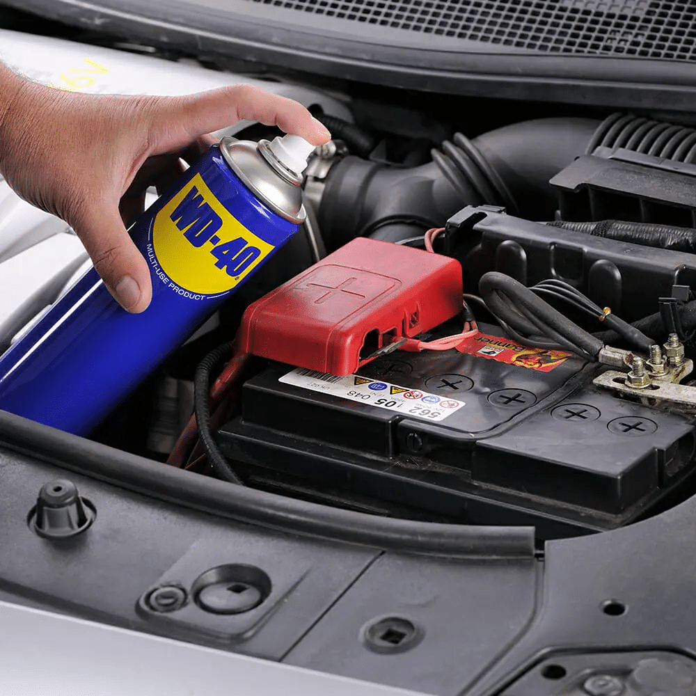 Primer plano del compartimento del motor de un coche con una mano sosteniendo un aerosol azul etiquetado como WD-40 Multiusos cerca de los terminales de la batería. La batería tiene un terminal positivo (+) rojo y un terminal negativo (-) negro visibles. La imagen sugiere el uso de WD-40 Multiusos ¿Para qué sirve? para el mantenimiento en las conexiones eléctricas de la batería del coche.