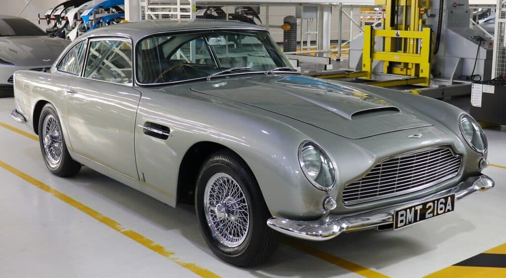 La imagen muestra un Aston Martin DB5 clásico de color plateado, con la matrícula “BMT 216A”, estacionado dentro de lo que parece ser un garaje o sala de exposición. Este vehículo es conocido por su asociación con la serie de películas de James Bond, lo que lo hace interesante y relevante para los fanáticos de la franquicia y los entusiastas de los coches clásicos.