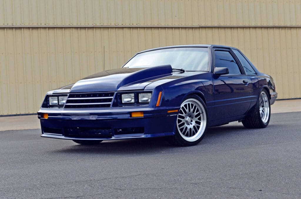Imagen de un Ford Mustang Foxbody de 1987 en color azul. El vehículo está estacionado con el frente ligeramente girado hacia la izquierda, mostrando su perfil lateral derecho. Cuenta con rines de aleación de múltiples radios y una carrocería bien cuidada que refleja la luz del entorno. No hay personas visibles ni otros objetos que distraigan en la imagen, lo cual pone todo el enfoque en el automóvil clásico.