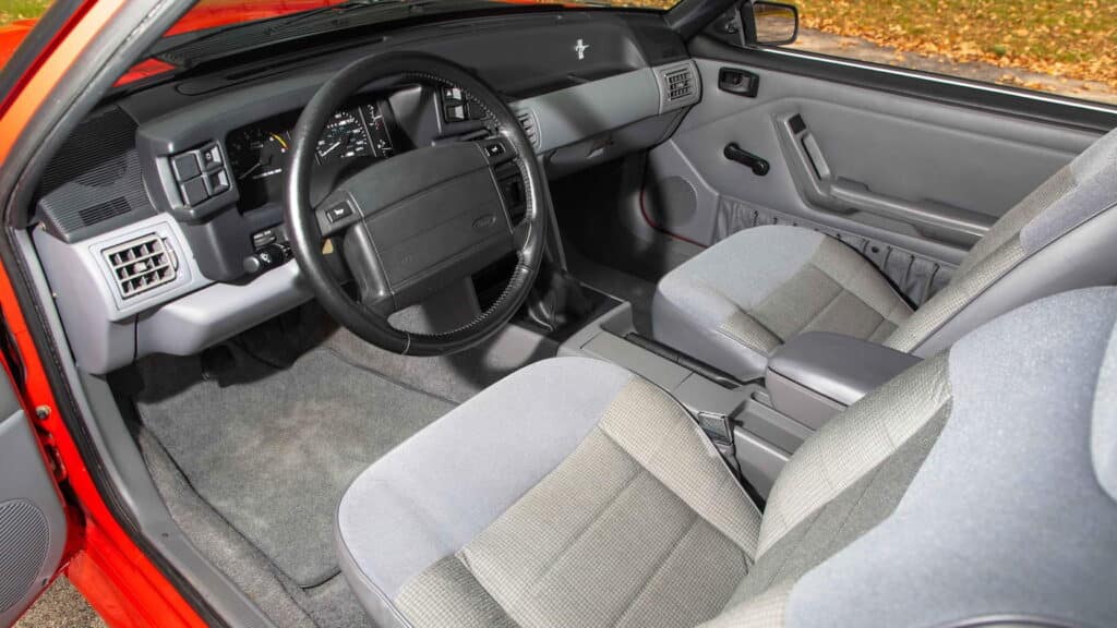 Vista interior de un Mustang Foxbody SVT mostrando dos asientos delanteros con tapicería gris, un tablero negro con varios controles y el icónico logo de Mustang en el volante. El vehículo tiene transmisión manual, evidente por la palanca de cambios en la consola central. La alfombra es gris, a juego con los asientos, y hay una rejilla de ventilación a la izquierda del volante. El panel de la puerta también es negro con controles de ventanas eléctricas.