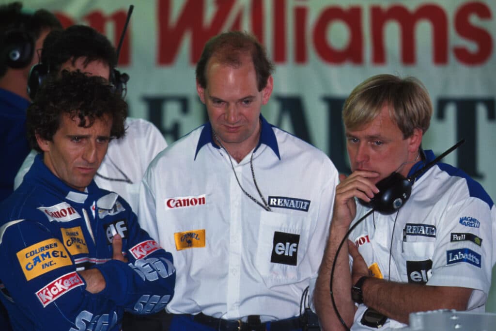 La imagen muestra a tres personas en un evento relacionado con la Fórmula 1 en los años 90. A la izquierda está Alain Prost con una chaqueta azul con logotipos de patrocinadores como “Canon” y “Renault”. En el centro está Adrian Newey con una camisa blanca y logotipos de “Renault” y “elf”. A la derecha hay una persona rubia con una chaqueta azul similar y un auricular alrededor del cuello, con logotipos como “Labatts”. Esta imagen destaca la colaboración entre Newey y Prost, dos figuras influyentes en la historia de la Fórmula 1.