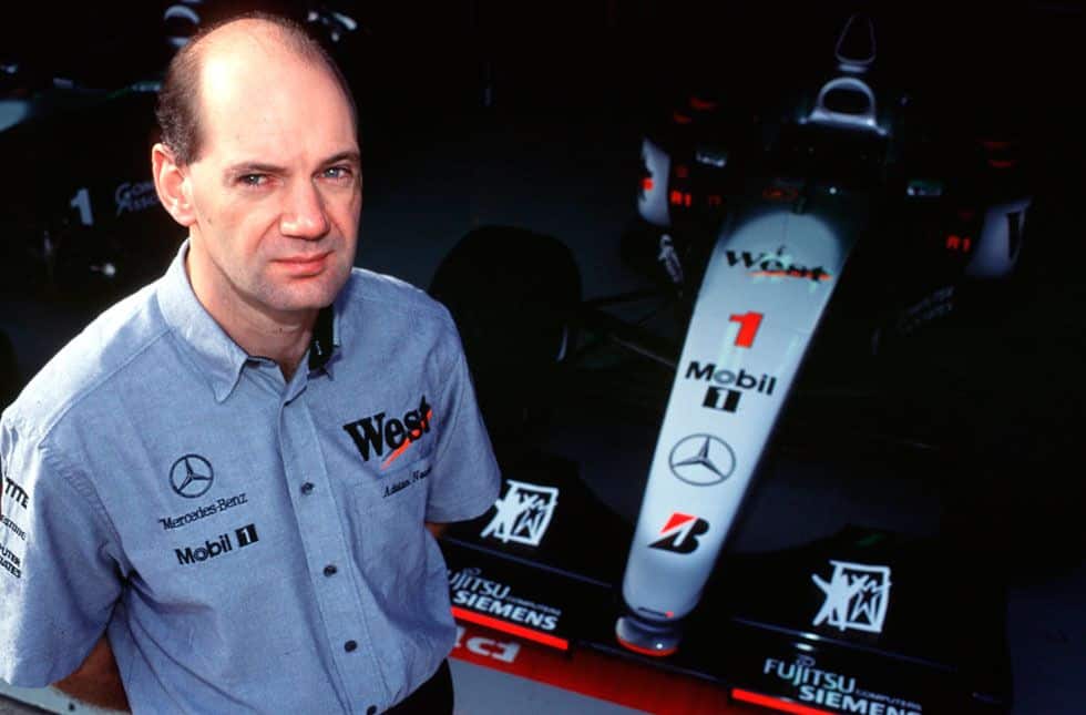 La imagen muestra a Adrian Newey junto a un coche de Fórmula 1 de McLaren en los años 90. El coche tiene varios logotipos de patrocinadores, incluyendo “West”, “Mobil 1” y el emblema de Mercedes. Esta imagen es relevante debido a la asociación de Newey con la Fórmula 1, un deporte conocido por su velocidad, tecnología y competencia global.