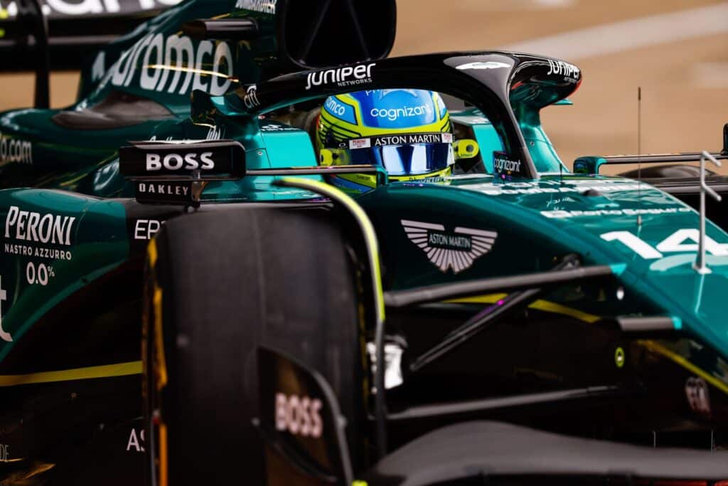 La imagen muestra un primer plano de un coche de Fórmula 1 del equipo Aston Martin. Se pueden ver elementos visibles como parte de la carrocería verde con logotipos de patrocinadores, el neumático Pirelli y una parte de la cabina con un piloto que lleva un casco azul con detalles amarillos. La carrocería presenta varios tonos de verde y elementos aerodinámicos como alerones y tomas de aire. Esta imagen es relevante para aquellos interesados en el automovilismo, las carreras de Fórmula 1 o los detalles de diseño y patrocinio de vehículos de alto rendimiento.