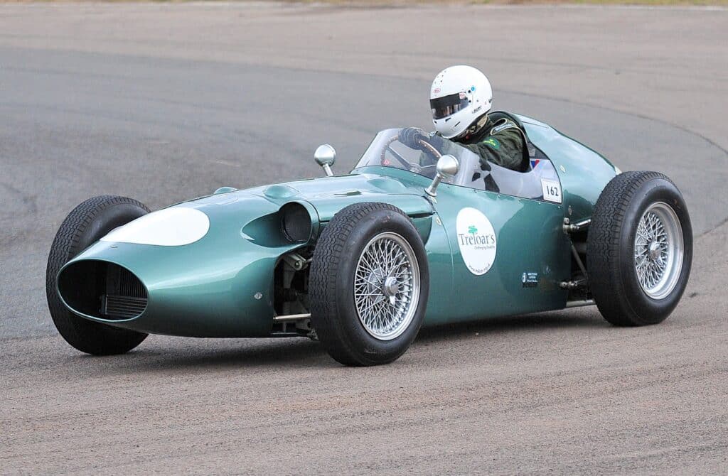 La imagen muestra un Aston Martin DBR4, un coche de carreras vintage en lo que parece ser una pista. El coche está pintado de verde y tiene el número “156” en su lateral. Cuenta con grandes ruedas de radios, una cabina abierta con un solo asiento, y el conductor lleva un casco blanco. El vehículo se asemeja a los diseños de mediados del siglo XX, característicos de los coches de carreras clásicos de esa época.