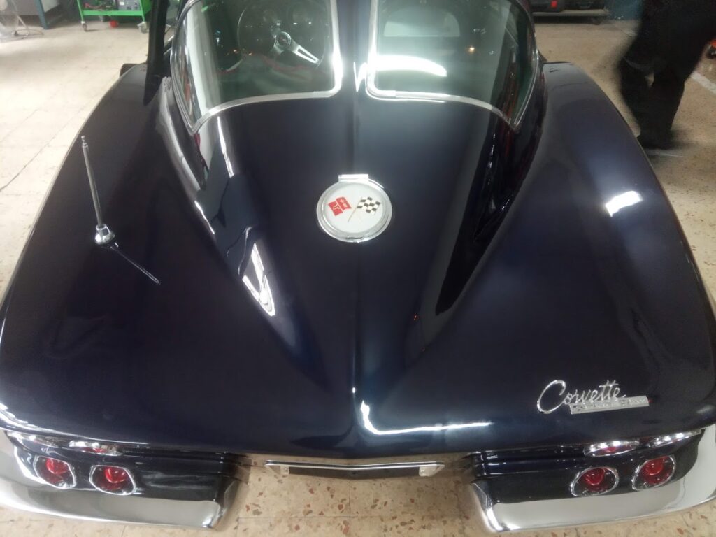 Vista trasera de un Chevrolet Corvette C2 Sting Ray de 1963, con su característica luneta trasera partida, estacionado en el taller de coches de lujo Jesauto en Zaragoza. El vehículo, de color rojo, muestra su diseño icónico y deportivo. En el fondo, se pueden ver herramientas y armarios de almacenamiento.