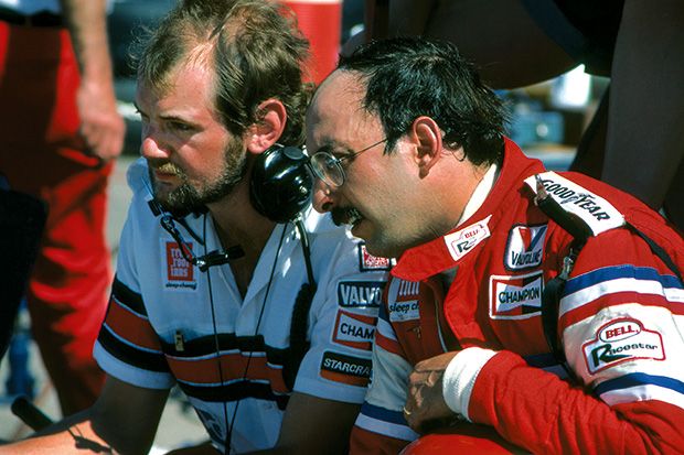 La imagen muestra a un joven Adrian Newey con barba en un entorno de boxes de carreras. Newey, a la izquierda, lleva una camisa blanca con detalles rojos y logotipos de patrocinadores. A su lado, una persona con un mono de carreras rojo y blanco con logotipos de “Valvoline” y “Champion” y un auricular, sugiere su rol en la comunicación con el equipo.